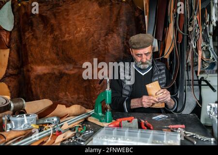 Uomo anziano che fa prodotti artigianali con vera pelle nella sua officina. Sfondo in pelle pelosa. Foto Stock