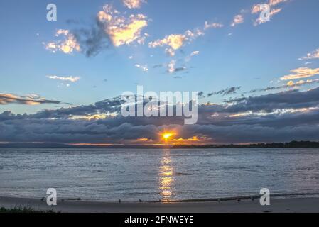 Uccello sulla riva guardando il sole che scende sul acqua e sbirciando tra le nuvole scure vicino all'orizzonte con la luce che brilla sulle nuvole più in alto Foto Stock