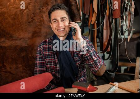 Giovane artigiano seduto sulla scrivania della sua officina artigianale che lavora con vera pelle mentre parla al telefono con viso sorridente. Foto Stock