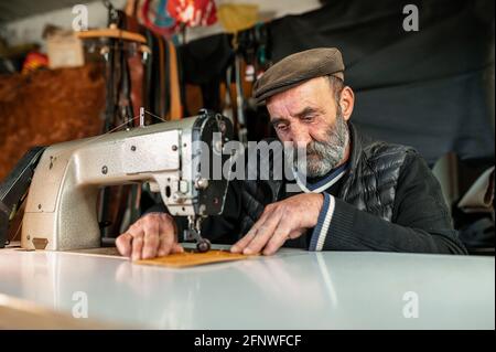 Vecchio uomo con bearded hippster con cappuccio piatto che lavora con pelle usando una macchina da cucire. Concetto artigianale. Foto Stock