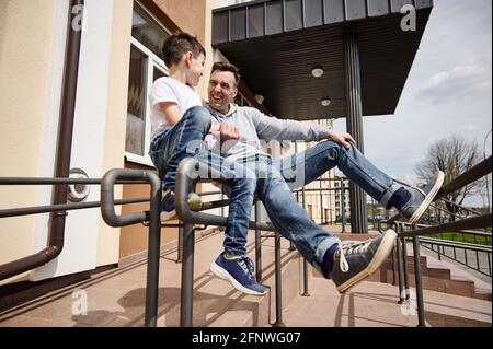 Bell'uomo caucasico che ride con il suo adorabile figlio mentre si siede su corrimano all'aperto Foto Stock