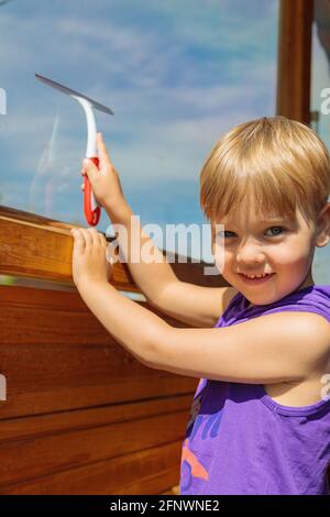 Il bambino lava i finestrini con una spazzola speciale. Un bambino con capelli biondi. Foto Stock