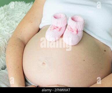 Closeup di donna incinta che tiene in mano le stivalette rosa a maglia del bambino sopra il ventre Foto Stock