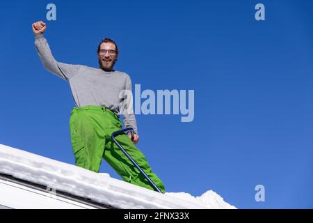 Divertente ritratto dal basso di un giovane uomo in piedi sul tetto di una casa con il braccio sollevato e una pala blu, durante un'operazione di rimozione della neve. Foto Stock