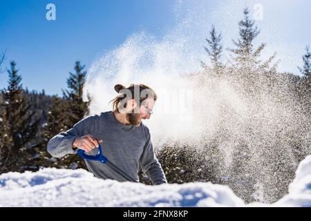 Vista laterale di un giovane che lancia energicamente un mucchio di neve fresca, creando una bizzarda, utilizzando una pala blu in una soleggiata giornata invernale canadese. Foto Stock