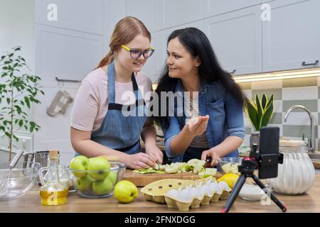 Mamma e figlia teen cucinano insieme la torta di mele, guardando lo schermo dello smartphone Foto Stock