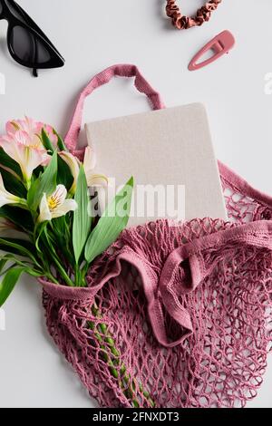 Fiori e taccuino di carta in borsa a corda su sfondo bianco. Elegante borsa da shopping femminile con vista dall'alto. Foto Stock