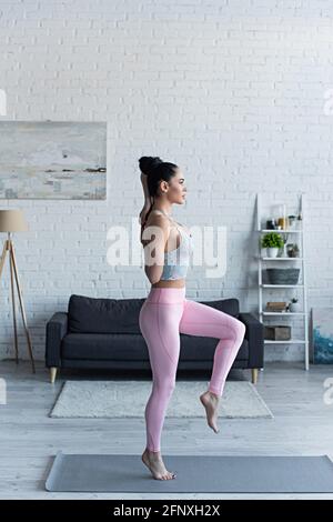 vista laterale di giovane donna in piedi su una gamba con mani dietro la schiena mentre si pratica yoga a casa Foto Stock