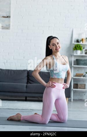 giovane donna a piedi nudi in abbigliamento sportivo in piedi sulle ginocchia sullo yoga stuoia e sorridente alla telecamera Foto Stock