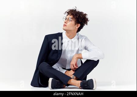 un ragazzo dai capelli ricci in abiti e sneaker alla moda si siede sul pavimento in una stanza luminosa in piena crescita Foto Stock