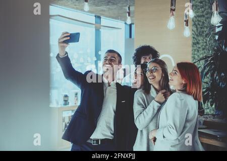 Gruppo di allegri colleghi che prendono selfie e gesturing mentre si levano in piedi nell'ufficio moderno. Foto Stock