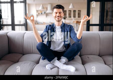 Meditazione, relax, riposo. Caucasica calma soddisfatto ragazzo in abiti casual elegante si siede sul divano nel soggiorno con gli occhi chiusi, medita in posizione lotus, allevia lo stress Foto Stock