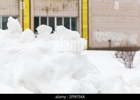 Una grande nevi bianca accatastata su una strada cittadina sullo sfondo di una casa. Un blocco di neve con grandi grumi si trovano in città nel cortile di un edificio di appartamenti. Giornata invernale cupa. Foto Stock