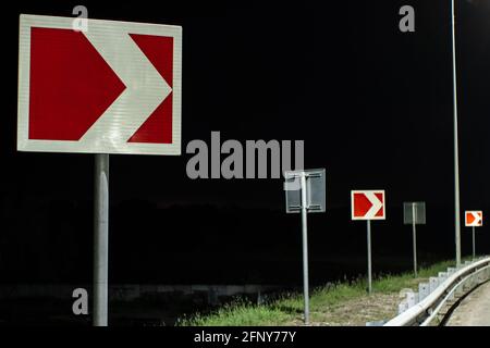Svolta a destra sulla strada notturna. Le indicazioni stradali segnalano curve strette alla luce dei lampioni Foto Stock