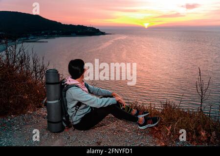 Una giovane donna con uno zaino sulla schiena, seduta sul bordo di una scogliera e ammirando il tramonto. Sullo sfondo, il mare, la montagna e lo sk Foto Stock