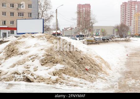 Un mucchio grande di neve sporca sul lato della strada con le automobili che passano. Sullo sfondo vi sono alti edifici di una moderna metropoli. Sullo sfondo di un cielo nuvoloso grigio. Foto Stock