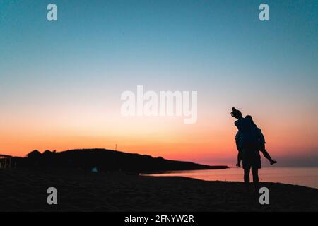 Silhouette di bella coppia sulla spiaggia al tramonto. Donna che cavalcava o si siede sulle spalle dell'uomo Foto Stock