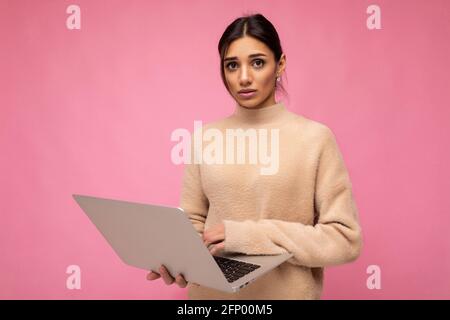 Foto di bella triste cupa giovane bruna donna che indossa un pullover beige tenendo netbook computer digitando testo sulla tastiera guardando la fotocamera isolata Foto Stock