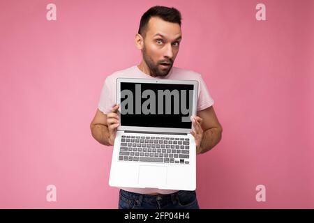 Bell'uomo sorpreso e stupito che tiene un computer portatile guardando fotocamera in t-shirt su sfondo rosa isolato Foto Stock