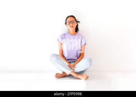 Corpo pieno di positivo a piedi nudi millennial asiatico femmina in casual camicia e jeans seduti sul pavimento in studio luce Foto Stock