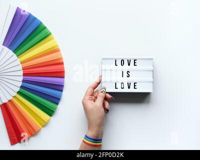 LGBTQ orgoglio mese decorazione. Testo Love is Love on lightbox. Ventola Rainbow pattern su sfondo bianco spento. La mano umana con vernice per unghie arcobaleno tiene la luce Foto Stock