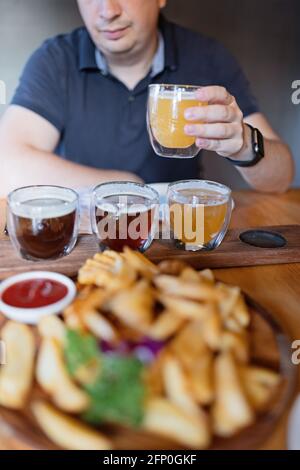 Uomo assaggiando varietà di birre artigianali stagionali in pub. Campionatori di birra in piccoli bicchieri posizionati singolarmente in fori realizzati in un unico vassoio di legno Foto Stock