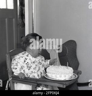 1961, storico, buon compleanno, con la sua bocca aperta, un bambino neonato seduto nella sua sedia in legno alto incerto di cosa a questo con la sua torta di compleanno e solitaria candela non illuminata, Inghilterra, UK. Foto Stock