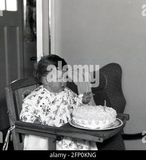 1961, storico, un bambino che si siede nella sua seggiolone di legno con la sua torta di compleanno Foto Stock