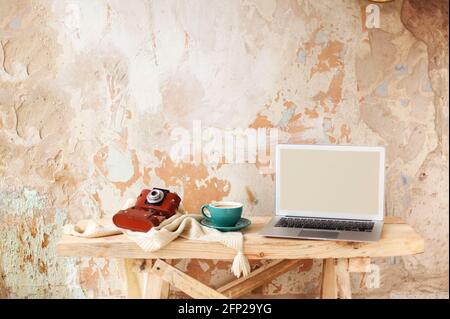 Fotocamera retrò e tazza di caffè fresco maglieria vicino a netbook moderno con schermo vuoto su tavolo di legno contro parete grugnosa di gesso in cre Foto Stock