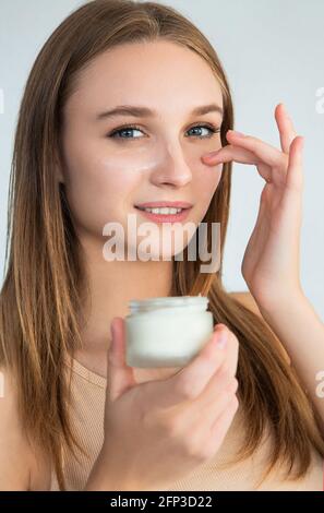 Giovane donna sorridente e guardando la macchina fotografica mentre applica idratante crema sul viso su sfondo blu Foto Stock