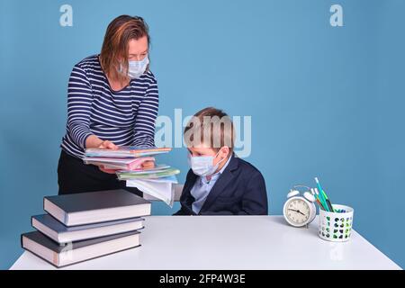 Mamma e figlio al banco della scuola che fanno i compiti nelle maschere mediche, sfondo blu Foto Stock