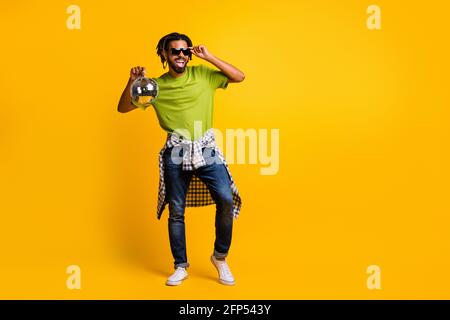 Foto di giovane uomo godere festa entusiasta Happy Hold discoteca pallina mano tocco occhiali da sole indossare jeans denim isolato sopra il giallo sfondo a colori Foto Stock