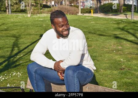 Felice uomo afroamericano seduto in un parco con jeans, maglione bianco. Concetto di stile di vita. Foto di alta qualità Foto Stock