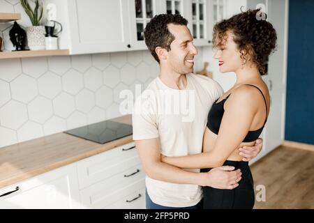 Felice giovane coppia stand in cucina moderna a casa hugging godersi la mattina insieme. L'uomo e la donna millennial sorridenti celebrano il trasferimento verso il nuovo Foto Stock