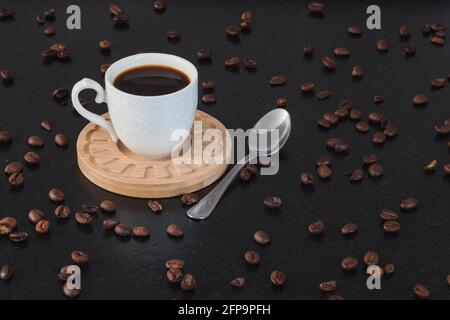 Una tazza bianca con caffè su un piatto di legno, con un cucchiaio di metallo su una panca di granito nero e molti chicchi di caffè intorno ad esso. Foto Stock