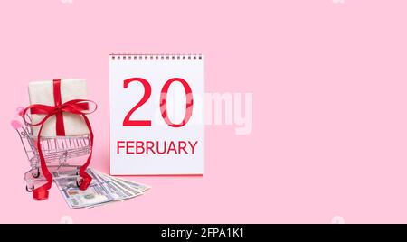 20 febbraio. Una confezione regalo in un carrello, dollari e un calendario con la data del 20 febbraio su sfondo rosa. Mese invernale, giorno di Foto Stock