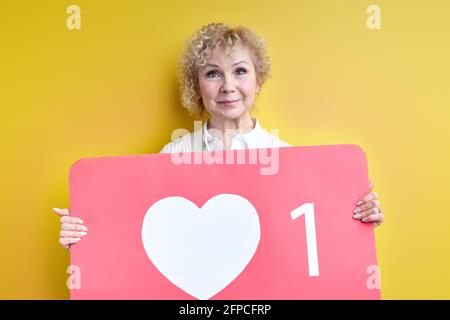 Donna anziano piacevole che tiene il cuore grande come segno, ottenga l'attenzione e nuovi seguaci in Blog, blogging nel concetto del Internet. Sfondo giallo isolato Foto Stock
