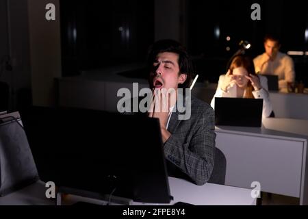 Uomo impegnato a urlare mentre si lavora su pc, in ufficio di notte. Ritratto di ragazzo in abbigliamento formale avendo scadenza, nessun tempo per dormire. Colleghi in th Foto Stock