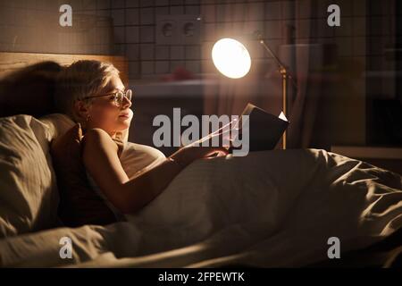 Bella femmina in pigiama e occhiali giacente sul letto di lettura libro, da sola di notte. Caucasica donna con capelli corti in camera da letto, bella donna in r Foto Stock