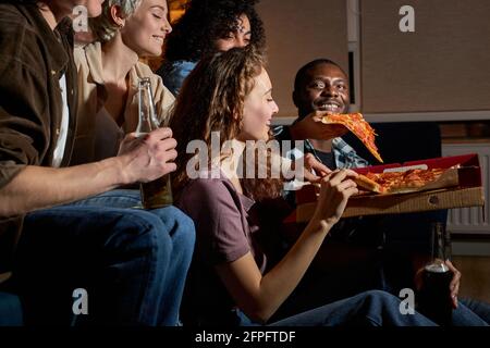 Gruppo di amici che mangiano pizza e guardano tv.Home party.concetto di fast food. Gli americani trascorrono del tempo libero insieme la sera. Vista laterale Foto Stock