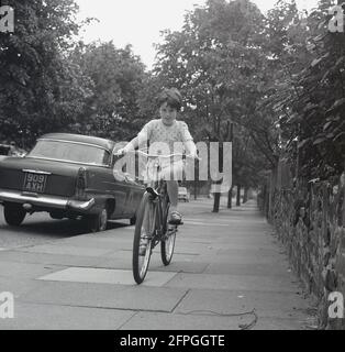 1969, storica, una giovane ragazza che guida la sua bicicletta con telaio in metallo, un Elswick Hopper 'Range Rider', sul marciapiede in una strada suburbana alberata, Inghilterra, Regno Unito. Una berlina dell'epoca, un Humber Super Snipe, è parcheggiata sulla strada. Foto Stock