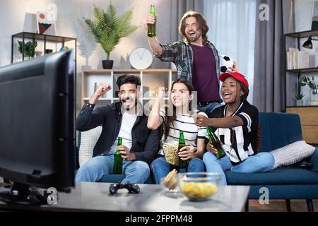 Gruppo di quattro amici multirazziali che gridano e che gestano mentre si guarda il campionato europeo di calcio in TV. I giovani fan sostengono emotivamente la squadra preferita da casa. Foto Stock