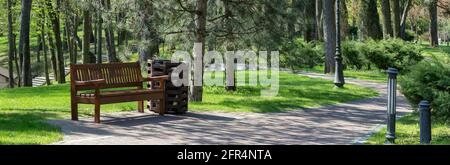 Parcheggia la panchina sotto un albero all'ombra. Bidone di rifiuti vicino alla panca. Sentiero per una passeggiata nel parco soleggiato in primavera Foto Stock