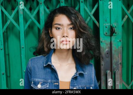 Headshot di una giovane bella ragazza asiatica in giacca blu jeans guardando la macchina fotografica, con uno sfondo verde. Foto Stock