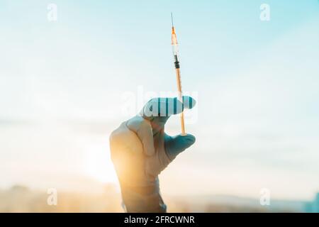 Siringa di vaccino del coronavirus tenuta dal medico o dall'infermiere in guanto blu medico dpi contro la riacutizzazione del tramonto. Immunizzazione che segna la fine della corona Foto Stock