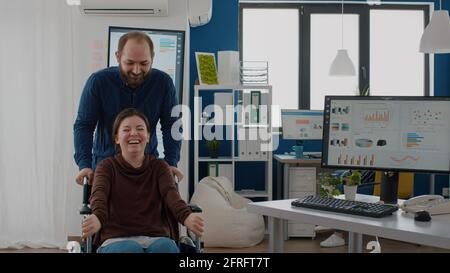Giovane uomo allegro che spinge la sedia a rotelle con il suo collaboratore invalido paralizzato mentre sorride in start up ufficio finanziario di affari. I colleghi si divertono a ridere sul posto di lavoro durante il lavoro Foto Stock