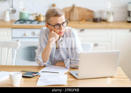 Frustrata donna matura indossare occhiali analizzare documenti e sensazione di triste, sconvolto, stanco a causa di alto costo di pagamento ipotecario. Lavorare su un computer portatile da casa. Persone anziane e concetto freelance Foto Stock
