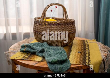 Interni vintage, calze a maglia e cesto di vimini con filato su un tavolo rotondo in legno coperto da una coperta folcloristica, primo piano Foto Stock