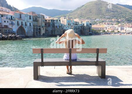 Bella giovane donna con cappello di paglia seduto su una panchina che gode di una vista del villaggio di Cefalu in Italia. Concetto di vacanze estive. Foto Stock