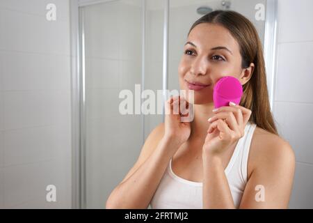 Giovane donna che pulisce la pelle del viso con speciale dispositivo a ultrasuoni durante la routine di igiene del mattino in bagno. Cura della pelle e concetto di benessere. Foto Stock